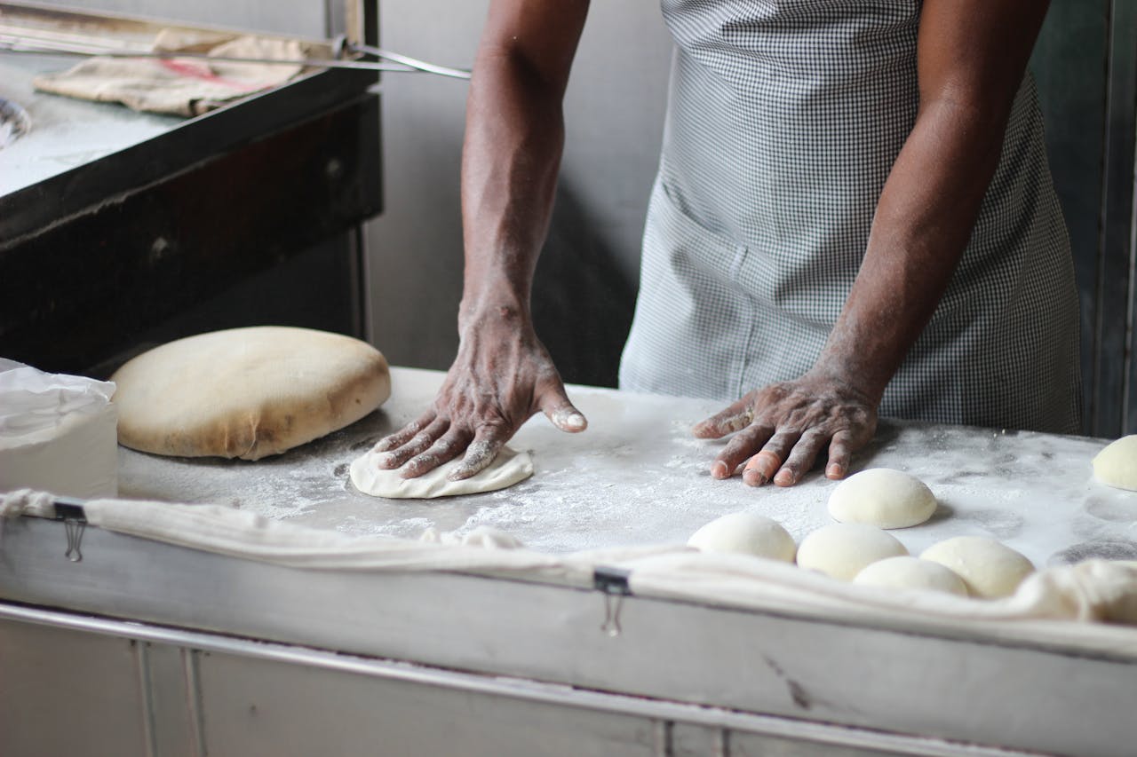 hero-img-02 A baker in an indoor kitchen kneading dough for bread preparation, showcasing traditional baking techniques.