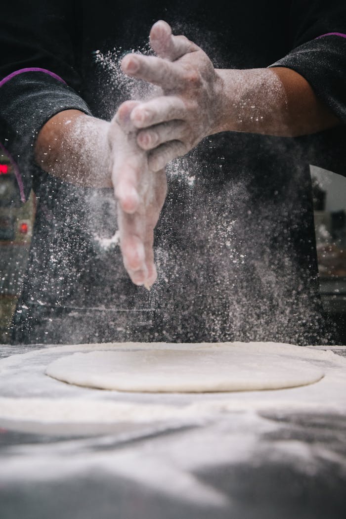gallery-2 A chef clapping hands over dough, releasing flour in a kitchen setting.